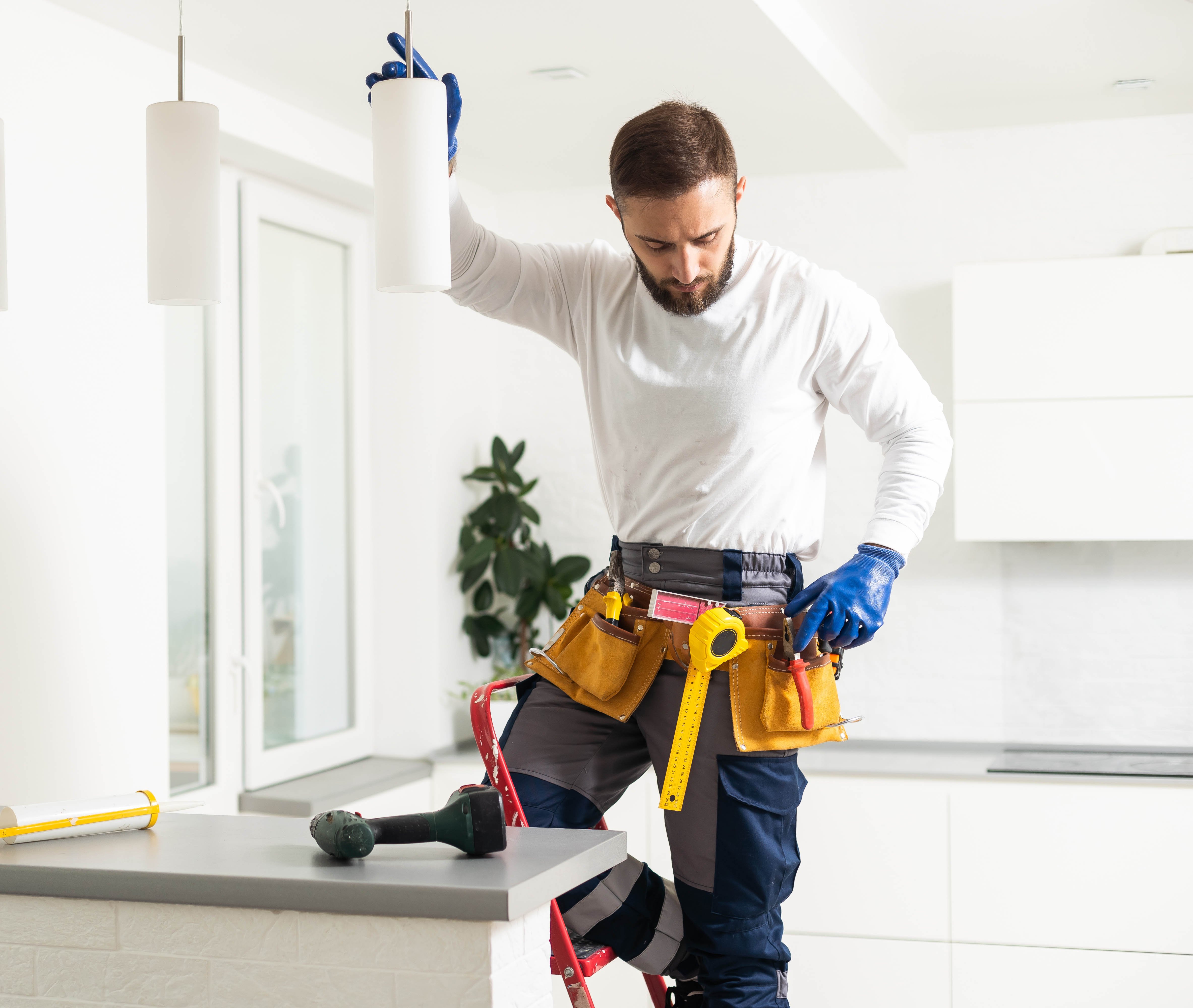 Tradesman in tool belt installing a ceiling light fixture in a modern kitchen