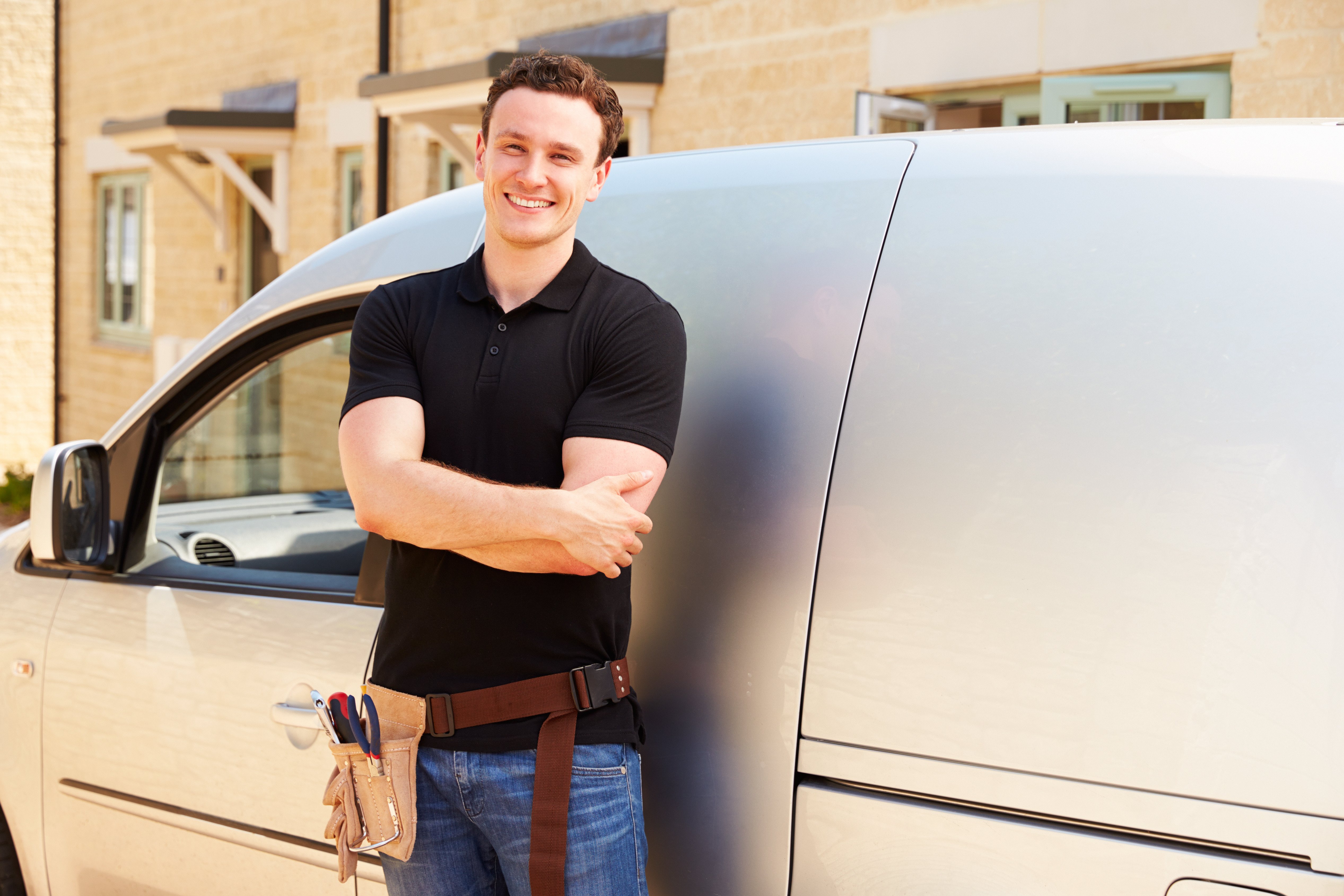 Smiling tradesman in a black shirt and jeans standing in front of his grey van with his arms crossed