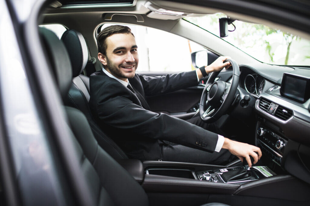 a smiling rideshare driver looks out of the passenger side door of a black vehicle