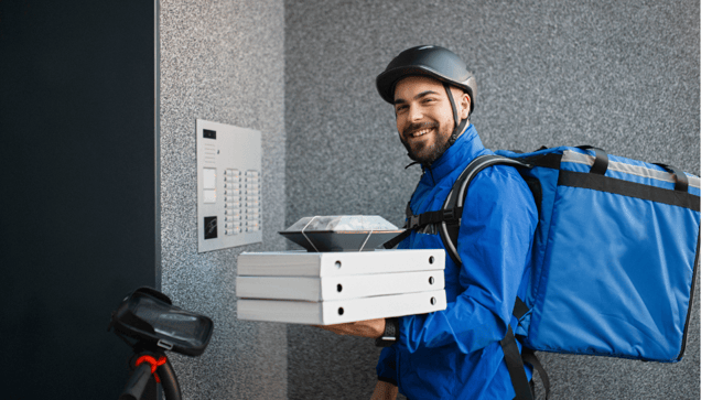 A food delivery courier dressed in blue smiling at a front door, holding a stack of pizza boxes