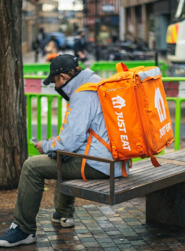 a just eat delivery driver looking at his phone. he is wearing a bright orange delivery rucksack