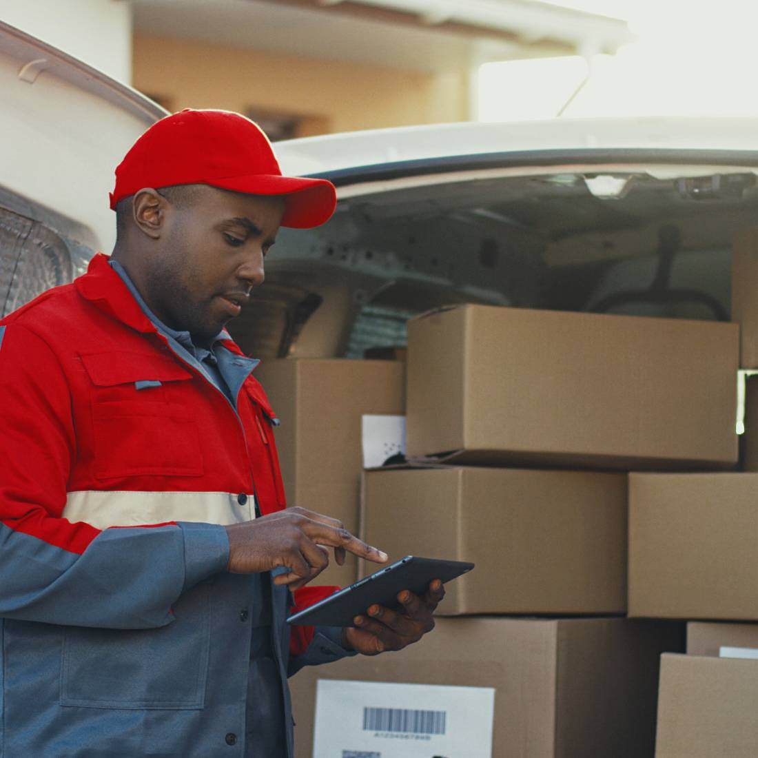 Delivery man with cap using a tablet next to a pile of boxes in his van