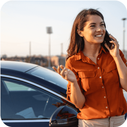 Woman in orange shirt leaning against her car and speaking on her phone