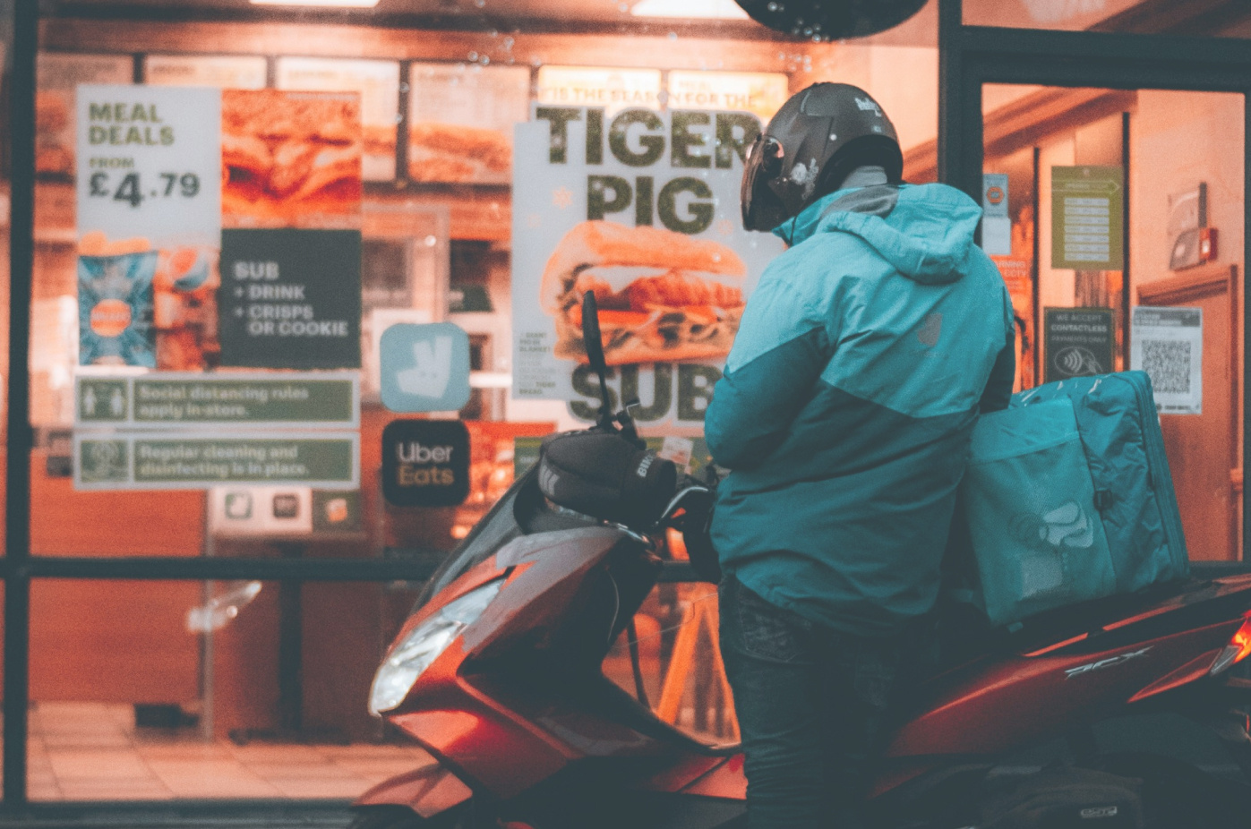 a fast food delivery driver waiting outside a restaurant. He is wearing a bright blue uniform, black motorcycle helmet.