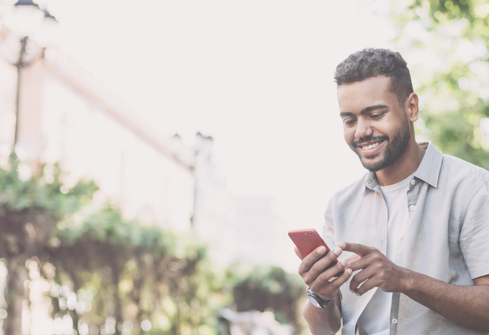 a man in a blue button up shirt holds his phone and is smiling. he is outside.