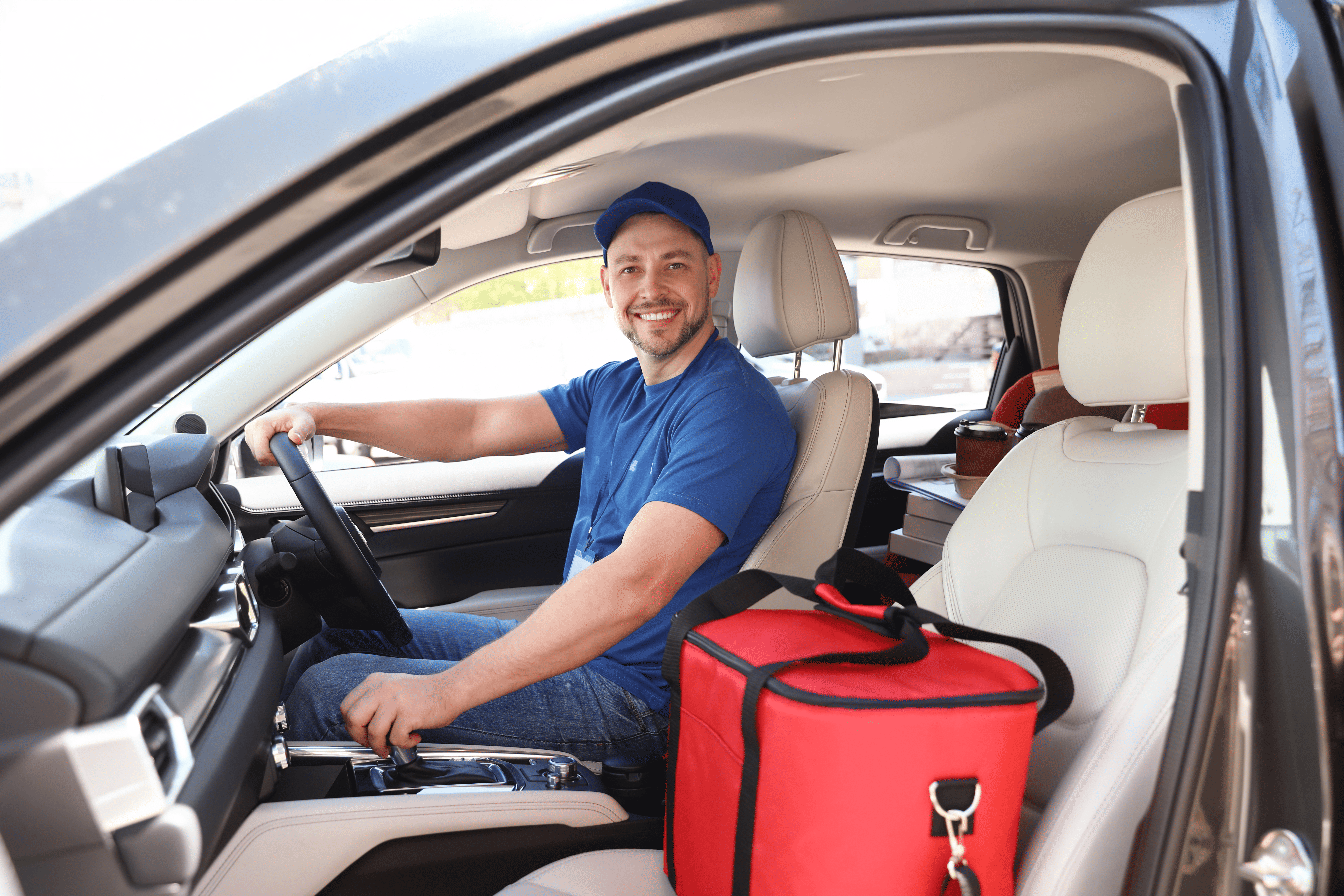 A professional delivery driver smiling in the driver's seat of a modern car, with a large red thermal food delivery bag and stacked parcel boxes on the front passenger seat.