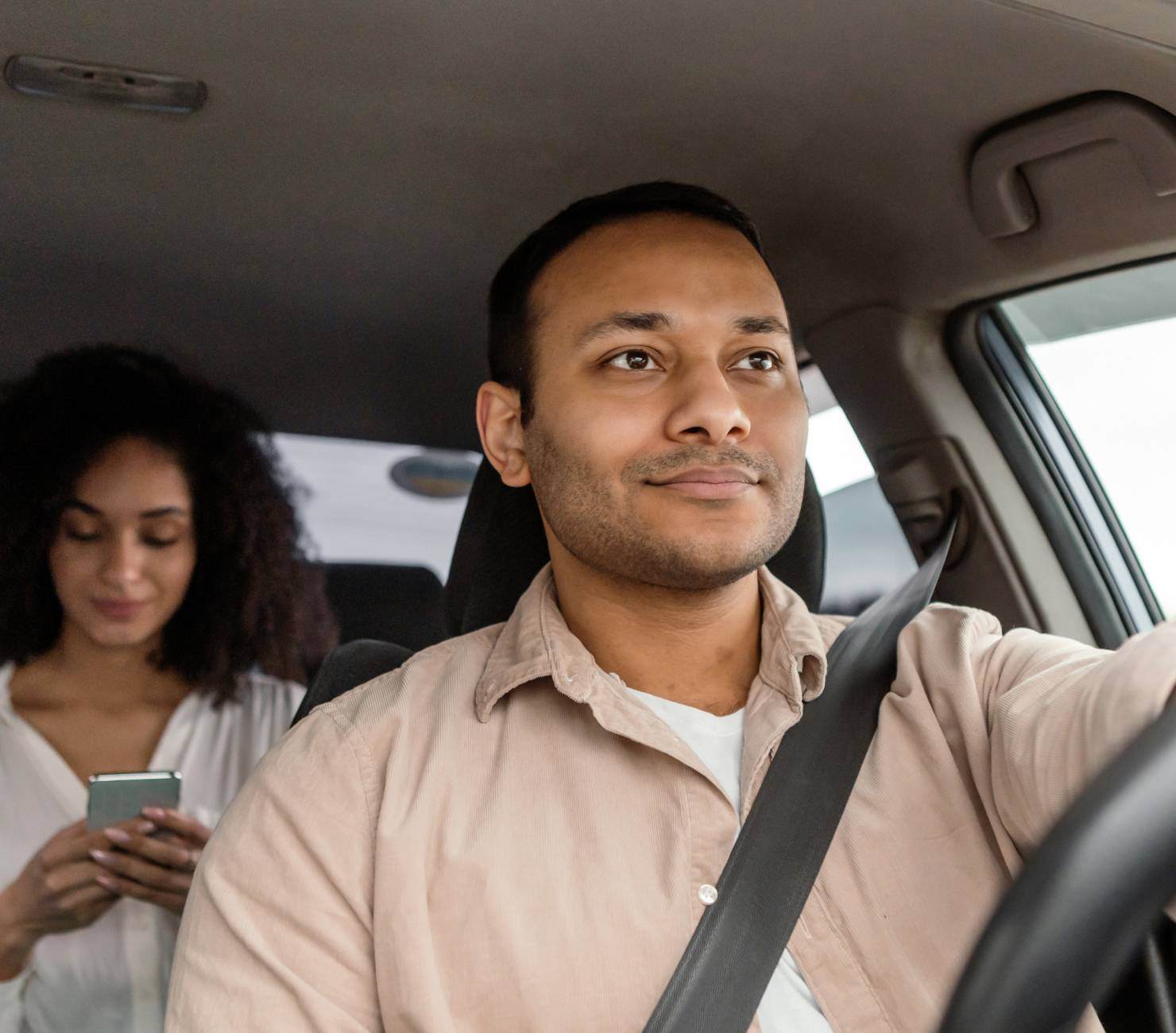 Smiling driver in a beige shirt and passenger who's looking at her phone