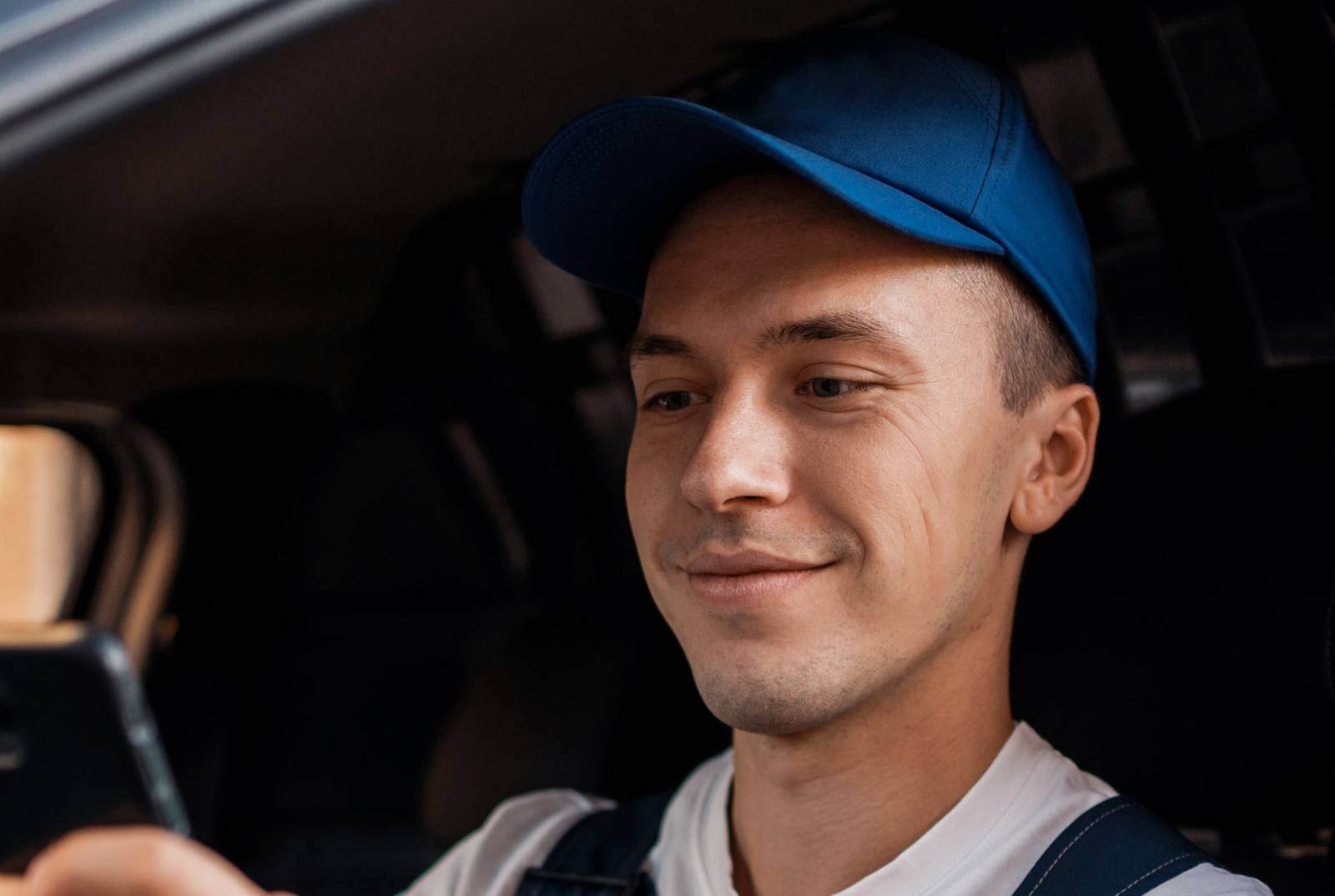 Driver in blue cap and overalls looking at his phone in his van