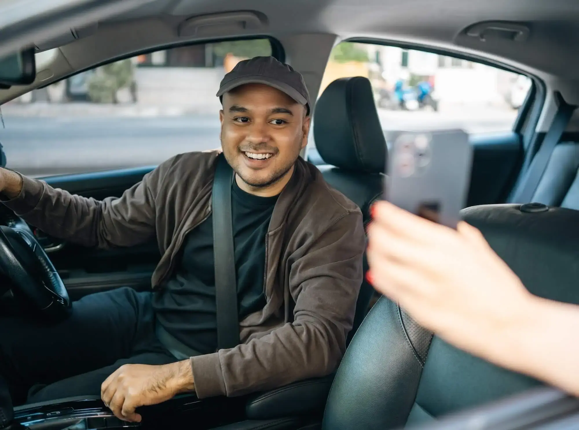 Driver in brown cap and jacket picking up a passenger and looking at her phone through the passenger-side window