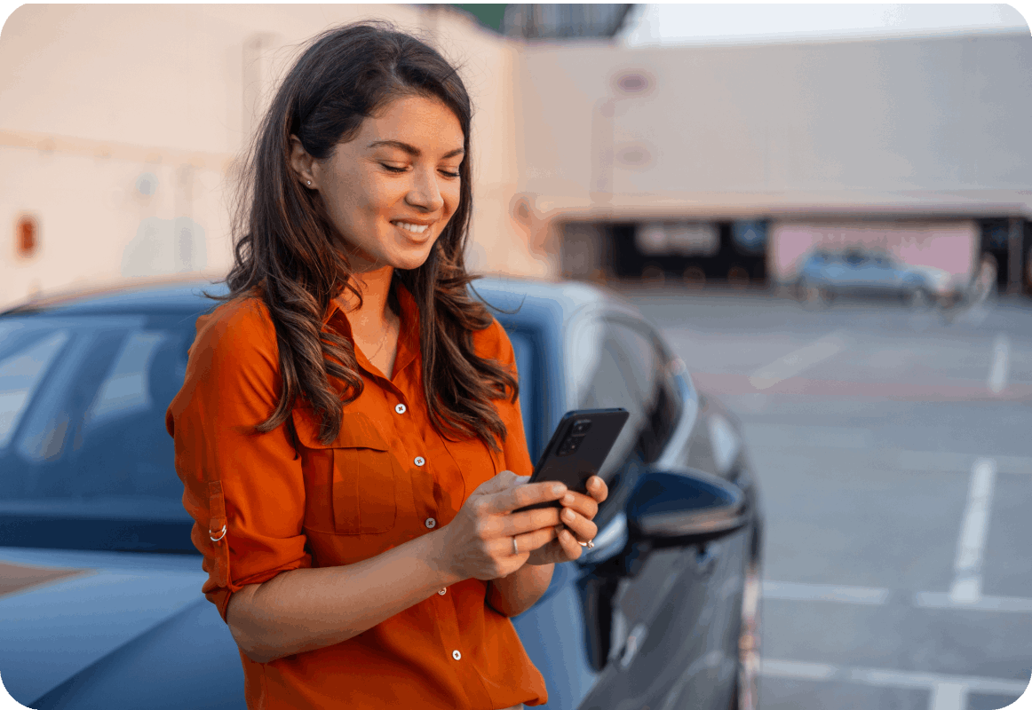 Woman in orange shirt leaning on the hood of her car, looking at her phone