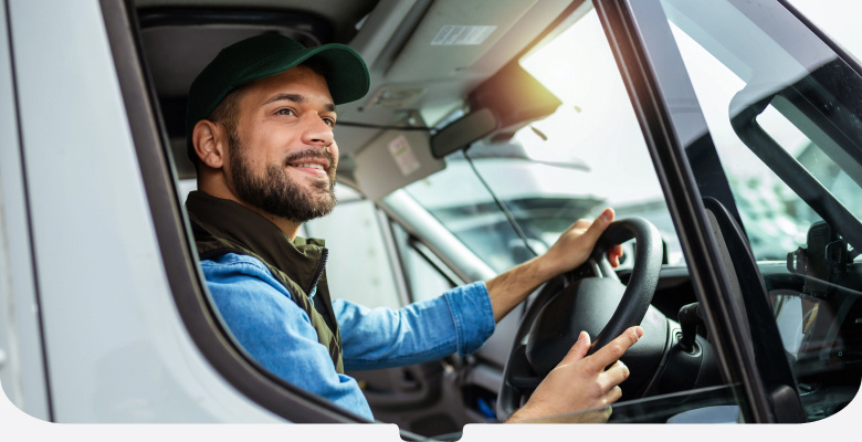 Bearded man in a blue denim shirt and black cap driving a van