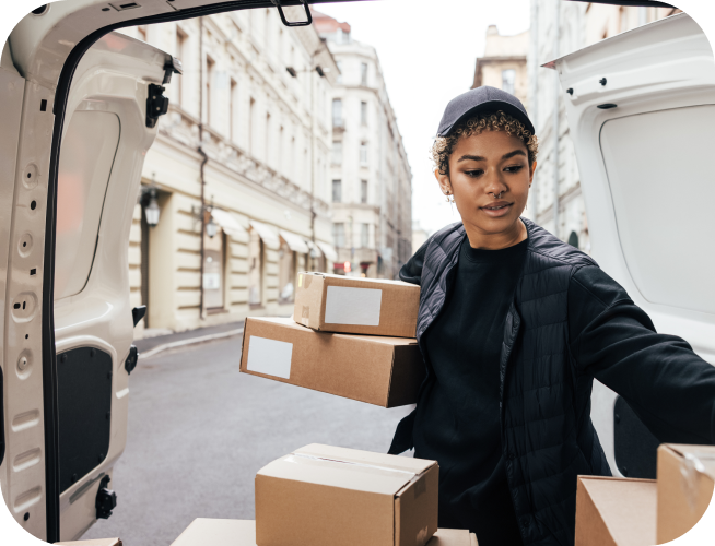 Woman with a nose ring, wearing a cap and black jacket, taking boxes out of a van