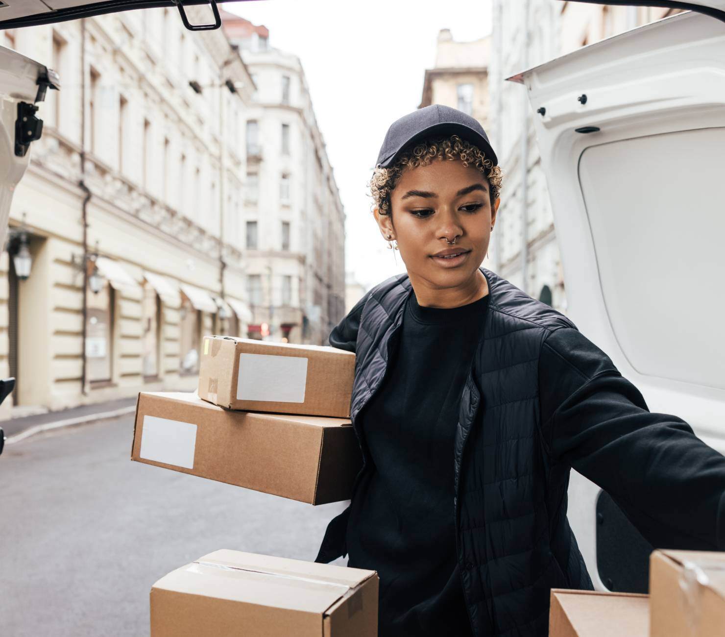 Woman with a nose ring, wearing a cap and black jacket, taking boxes out of a van