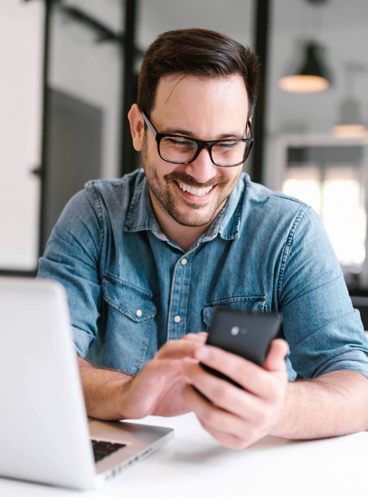 Smiling man in glasses and a blue denim shirt sitting in front of his laptop looking at his phone