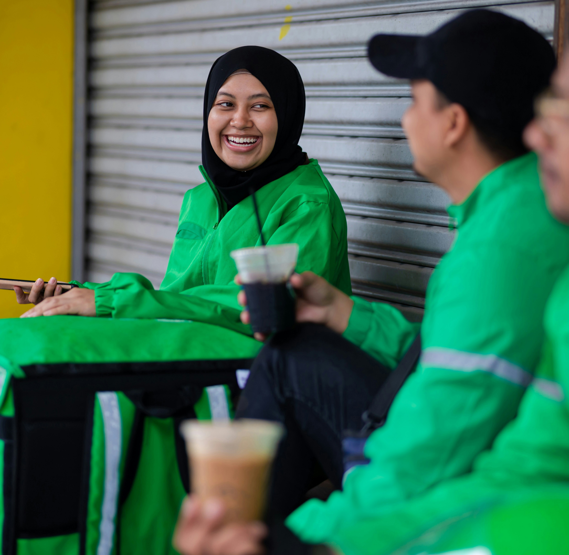 a female food delivery driver in a green uniform and black headscarf smiles at her colleagues, also wearing green