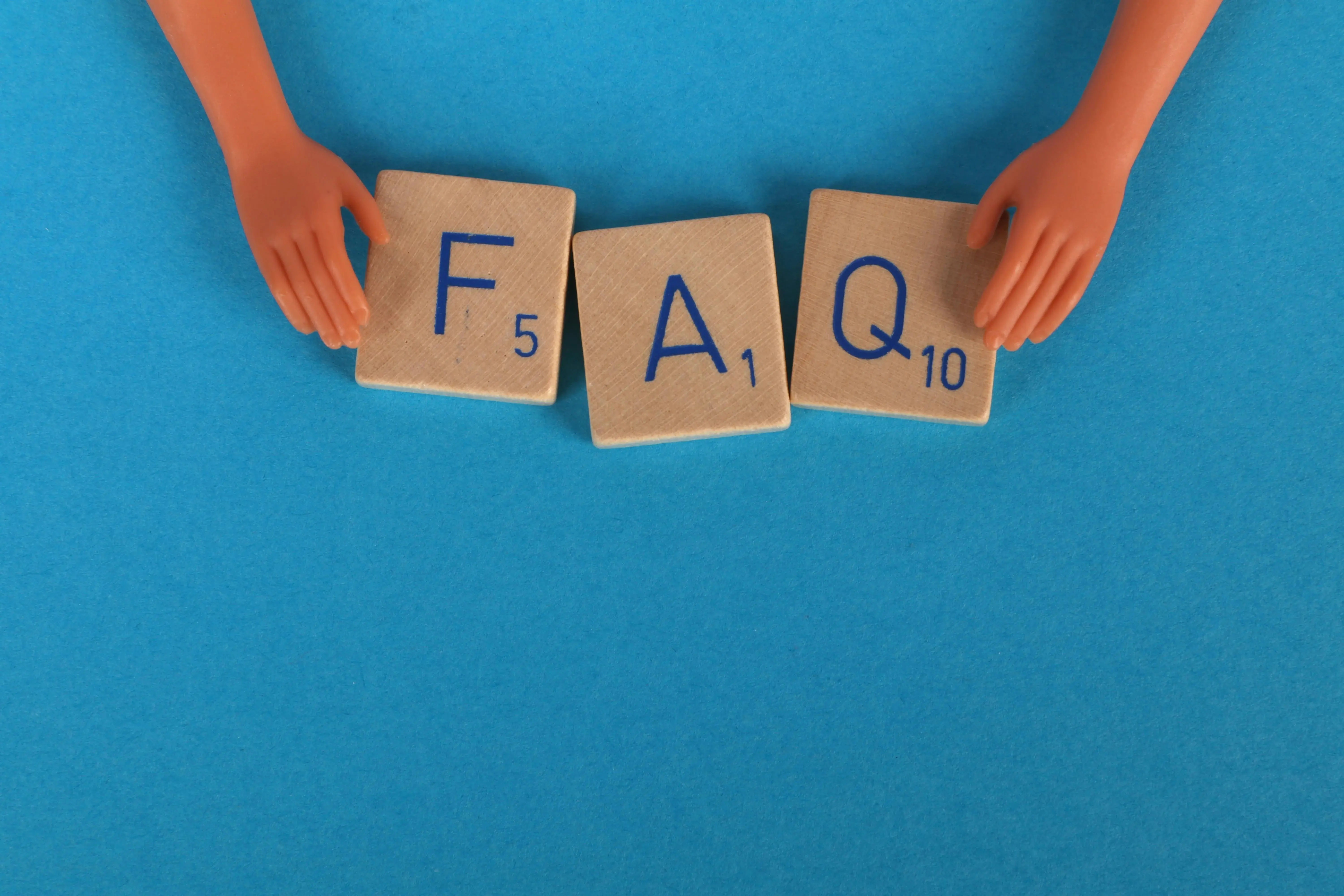 a pair of doll hands holding scrabble tiles that read F A Q on a blue background