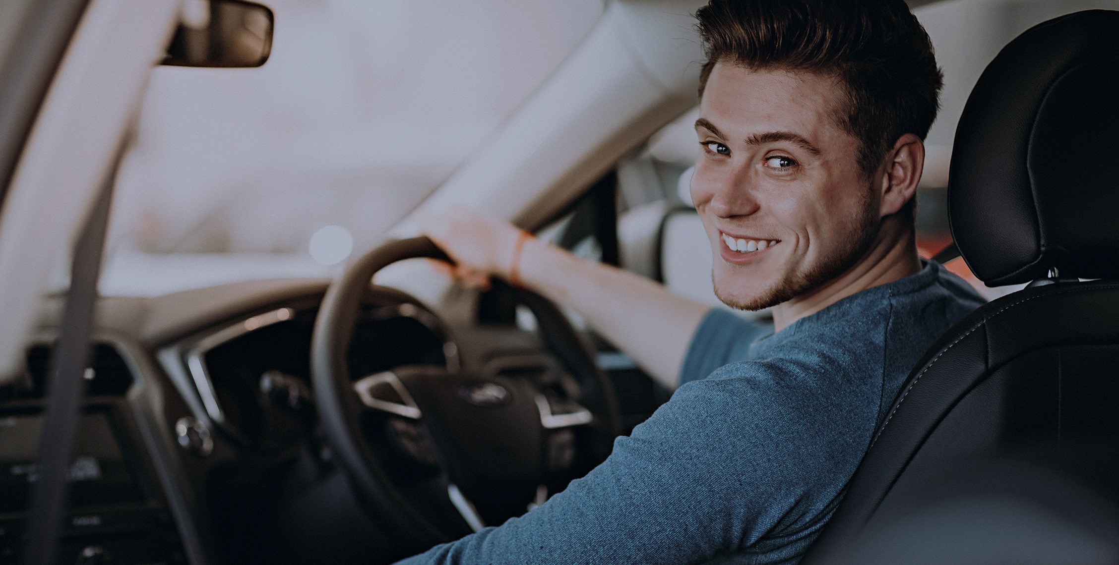 A young man in a blue shirt sat behind a steering wheel smiling to camera
