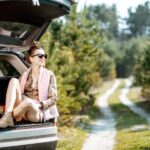 Young woman enjoying spring while sitting in the car trunk on a picturesque road in the woods